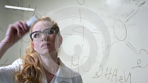 A Caucasian woman in a medical gown thinks and finalizes formulas on a transparent wall.