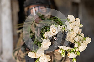 Caucasian woman in military uniform holding a machine gun and a bouquet of white roses.