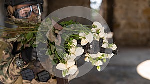 Caucasian woman in military uniform holding a machine gun and a bouquet of white roses.