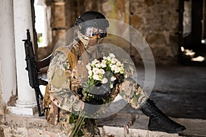 Caucasian woman in military uniform holding a machine gun and a bouquet of white roses.