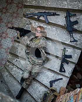 Caucasian woman in military uniform lies on the stairs of an abandoned building and holds a machine gun. View from above