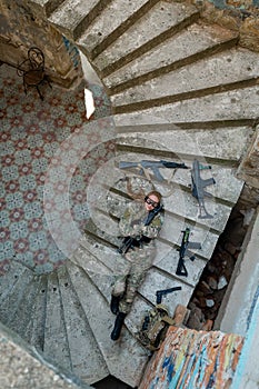 Caucasian woman in military uniform lies on the stairs of an abandoned building and holds a machine gun. View from above