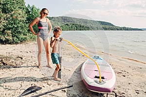 Caucasian woman mother teaching son to inflate sup surfboard with pump on beach.