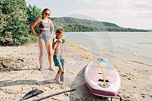Caucasian woman mother teaching son to inflate sup surfboard with pump on beach. photo