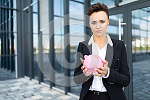 Caucasian woman in office clothes holds a pink pig moneybox and waits a coleague near the office building