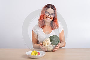 Caucasian woman prefers healthy food. Redhead girl chooses between broccoli and donuts on white background.