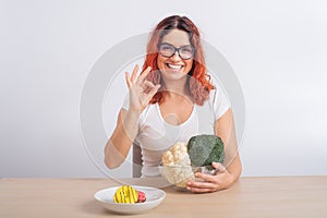 Caucasian woman prefers healthy food. Redhead girl chooses between broccoli and donuts on white background.