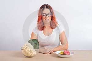 Caucasian woman prefers healthy food. Redhead girl chooses between broccoli and donuts on white background.