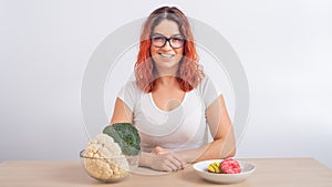 Caucasian woman prefers healthy food. Redhead girl chooses between broccoli and donuts on white background.