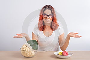 Caucasian woman prefers healthy food. Redhead girl chooses between broccoli and donuts on white background.