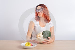 Caucasian woman prefers healthy food and refuses fast food. Redhead girl chooses between broccoli and donuts on white