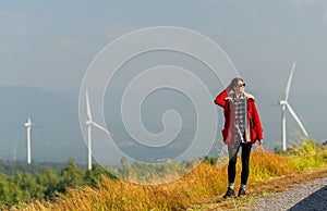 Caucasian woman with red coad and sunglasses walk along the roadside near the meadow and wind turbines or windmill also look to