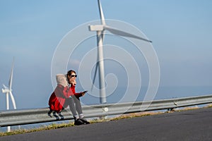 Caucasian woman with red coat and sunglasses sit on barrier of roadside and use mobile phone on mountain near windmill or wind