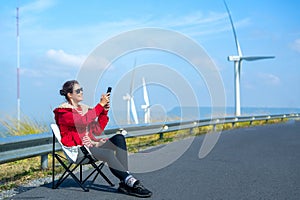 Caucasian woman with red coat and sunglasses sit on chair on roadside near wind turbines or windmill also use mobile phone to take