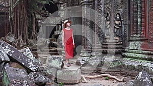Caucasian woman in red dress is walking among ruins of Ta Prohm