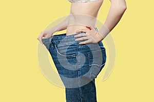 A Caucasian woman shows the results of her weight loss by pulling on wide-waisted blue jeans. Isolate, yellow background. Side