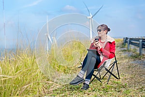 Caucasian woman sit on chair near meadow with blue sky and wind turbines or windmill on the back and she also hold a cup of coffee