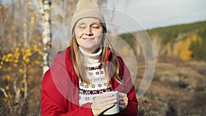 Caucasian woman is sitting alone in autumn forest, drinking hot tea from gray mug and enjoying nature. The wind gently