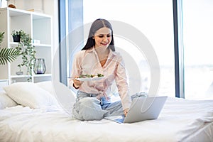 Caucasian woman sitting on comfy bed with wireless laptop, eating healthy salad