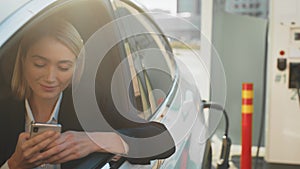 Caucasian woman sitting on driver seat in luxury car and smiling on camera through open window at EV station. Pleasant