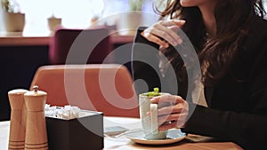 Caucasian woman sitting inside of cafe with a lot of green plants, drinking hot tea, touching her long brown hair. Close