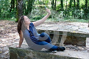Caucasian woman, with slightly curly hair, on top of a stone bench in a picnic area taking a selfie with her smartphone
