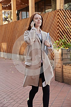 caucasian woman in stylish beige coat calling by smartphone, holding laptop