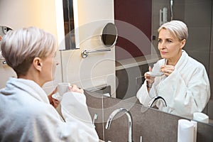 Caucasian woman with tea or coffee looking at herself in mirror in bathroom