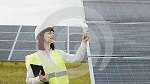 Caucasian woman in uniform and white helmet standing at solar farm and holding bulb. Female engineer using digital