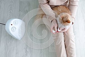 A caucasian woman warms herself with  fan heater, sitting on the floor in a room with a cat in her arms