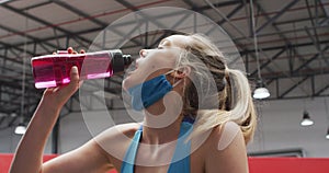 Caucasian woman wearing lowered face mask drinking water at gym