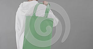 Caucasian woman wearing white hoodie holding green bag on grey background, copy space, slow motion