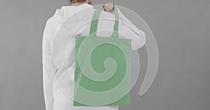 Caucasian woman wearing white hoodie holding green bag on grey background, copy space, slow motion