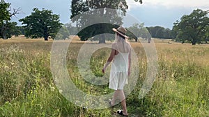 Caucasian woman with white dress walking a trail through a meadow with large oak trees during summer