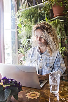 Caucasian woman working on laptop sitting at the desk with green plants in background. Concept of modern people and online