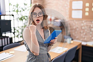 Caucasian woman working at the office wearing glasses surprised pointing with hand finger to the side, open mouth amazed