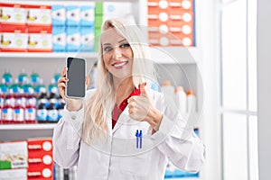 Caucasian woman working at pharmacy drugstore showing smartphone screen smiling happy and positive, thumb up doing excellent and