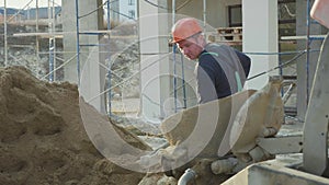 Caucasian worker pours sand into a concrete mixer by shovel at construction site