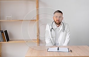 Caucasian young bearded male doctor sits in his office at the table with a tired look.