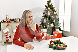 Caucasian young blonde woman sitting on the table by christmas tree bored yawning tired covering mouth with hand