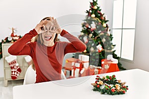Caucasian young blonde woman sitting on the table by christmas tree doing ok gesture like binoculars sticking tongue out, eyes