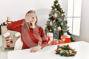 Caucasian young blonde woman sitting on the table by christmas tree peeking in shock covering face and eyes with hand, looking
