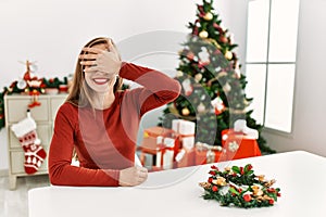 Caucasian young blonde woman sitting on the table by christmas tree smiling and laughing with hand on face covering eyes for