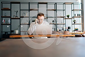 Caucasian young business man wearing fashion casual clothing working on laptop and talking on phone