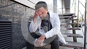 Caucasian young doctor man sits down on the stairs near the clinic building, tired and unhappy rubbing his nose and eyes