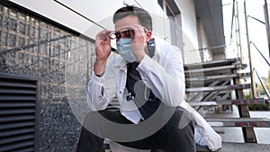 Caucasian young doctor man sits down on the stairs near the clinic building, tired and unhappy rubbing his nose and eyes