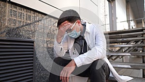 Caucasian young doctor man sits down on the stairs near the clinic building, tired and unhappy rubbing his nose and eyes