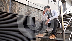 Caucasian young doctor man sits down on the stairs near the clinic building, tired and unhappy rubbing his nose and eyes