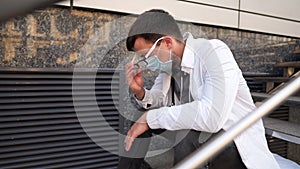 Caucasian young doctor man sits down on the stairs near the clinic building, tired and unhappy rubbing his nose and eyes