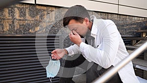 Caucasian young doctor man sits down on the stairs near the clinic building, tired and unhappy rubbing his nose and eyes